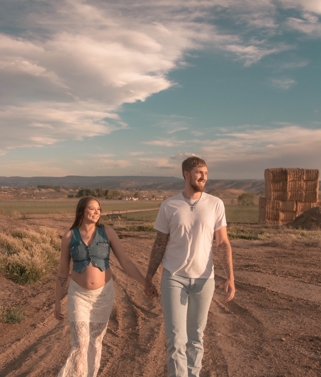 Ashley and Kevin walking hand in hand down a dirt road with the mesa and hay bales behind them