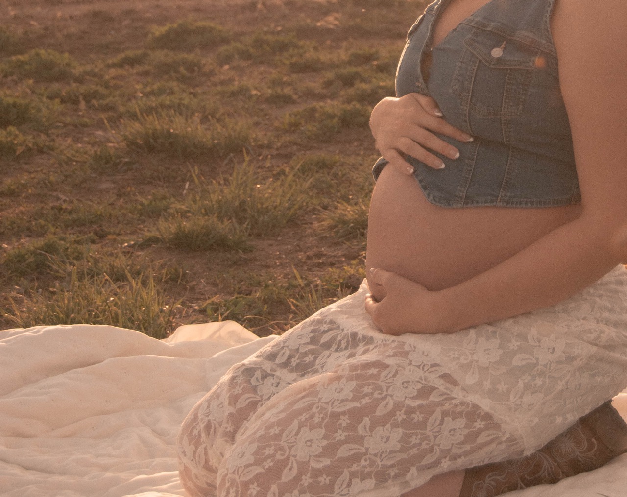 Ashley sitting on a quilt in a meadow at sunset cradling her belly with both hands