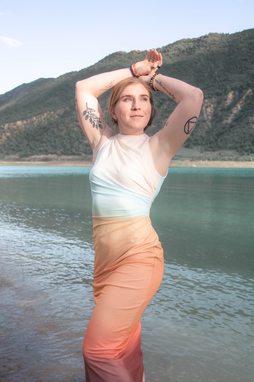 Woman in an ombre dress standing in a still lake with arms lifted overhead, mountain behind her