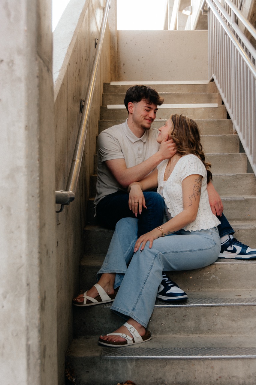 Ellie and Lane sitting on a sunlit concrete stairwell, foreheads close, smiling at each other