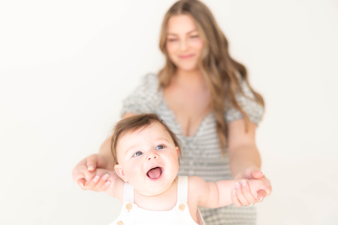 Smiling baby in white knit overalls reaching forward while mom in a sage gingham dress holds their hands in a bright studio