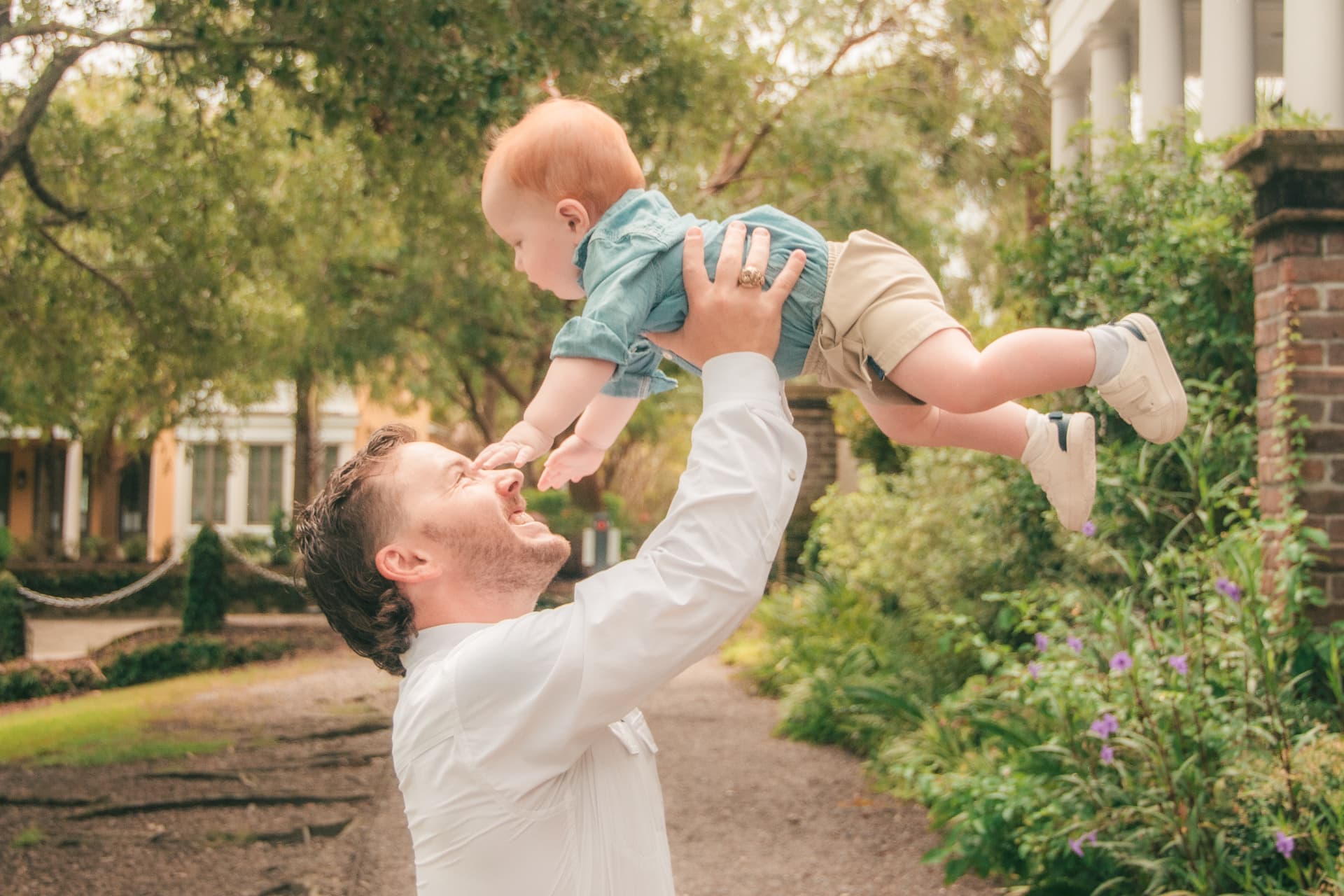 Dad lifting his baby boy high into the air beneath the trees, both of them laughing