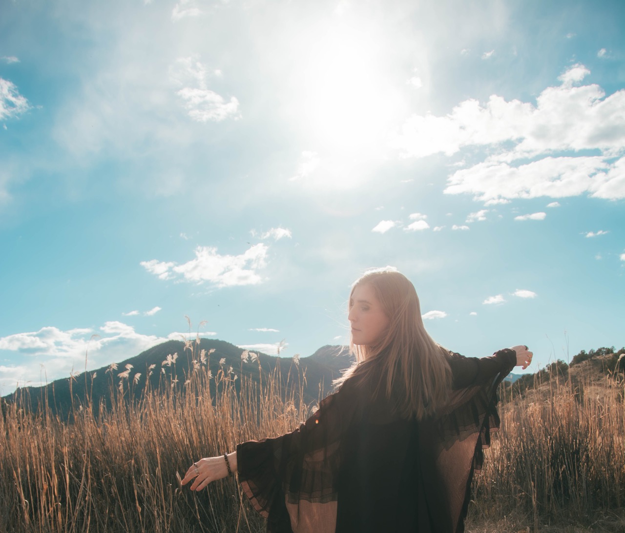 Woman in a black chiffon kimono with arms outstretched in a tall grass field, sun flaring behind her