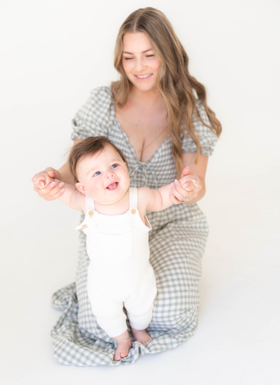 Baby standing barefoot on mom's gingham dress, laughing in a bright white studio