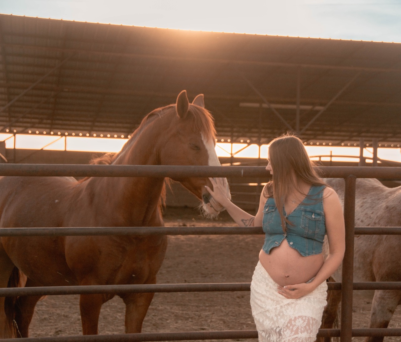 Ashley in a denim vest and lace skirt reaching up to greet a chestnut horse under the arena roof at sunset