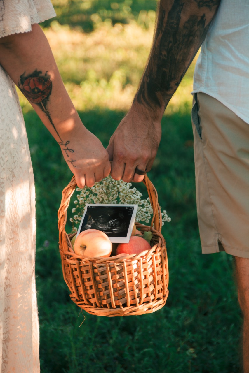 Couple carrying a picnic basket of apples, baby's breath, and their ultrasound photo through the orchard
