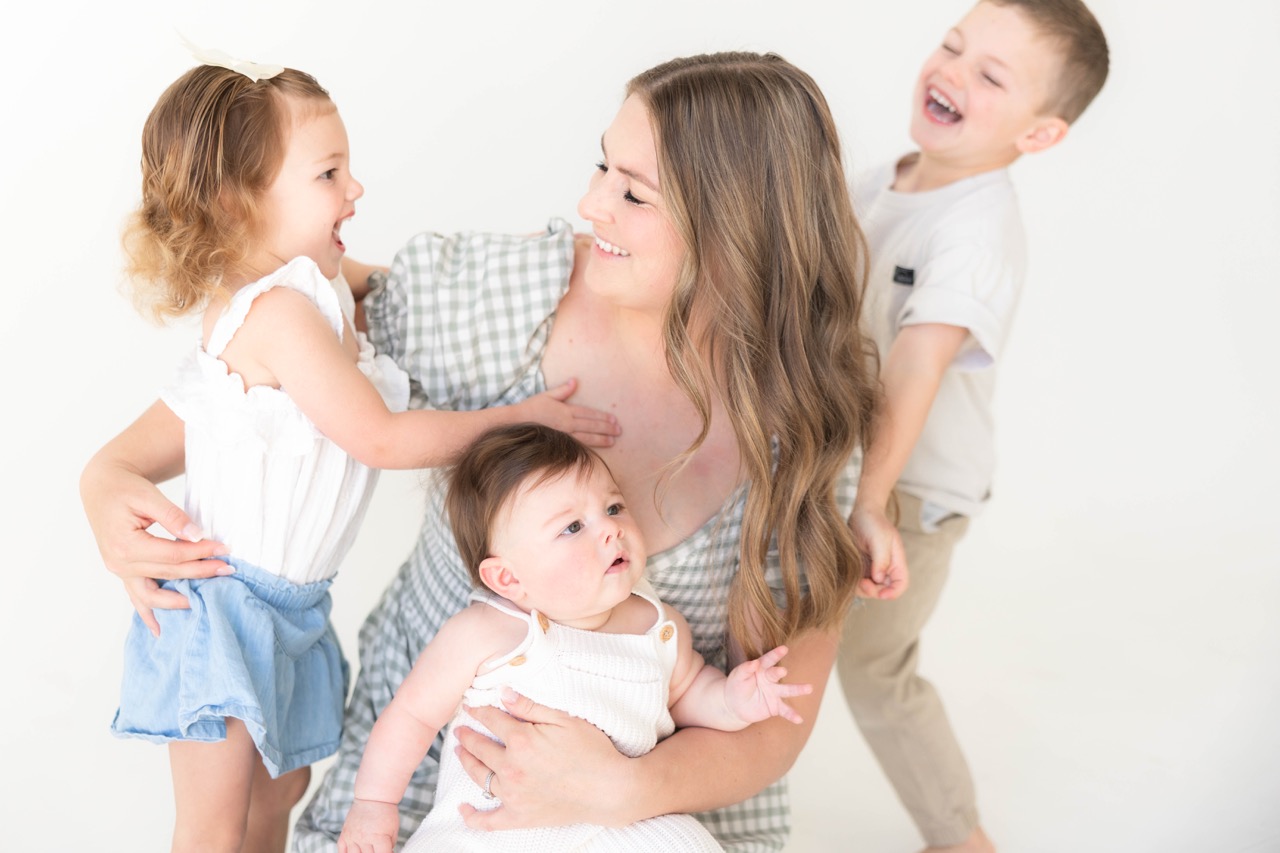 Three siblings laughing around their mom in a white studio — a toddler girl, a baby in her arms, and a big brother behind