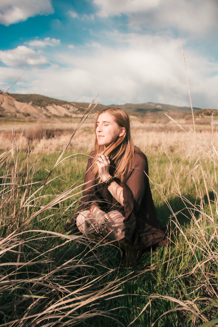 Woman in a brown chiffon dress kneeling in tall grass with eyes closed, soft afternoon light