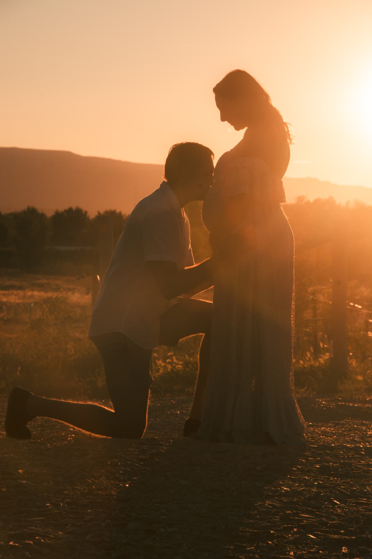 Sunset silhouette of a father-to-be kneeling to kiss his wife's pregnant belly