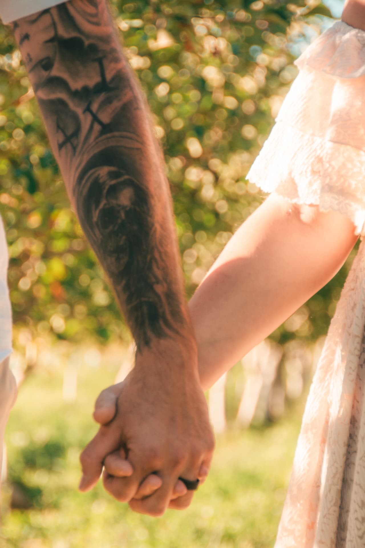 Close-up of a tattooed hand holding hers as they walk through the orchard grass