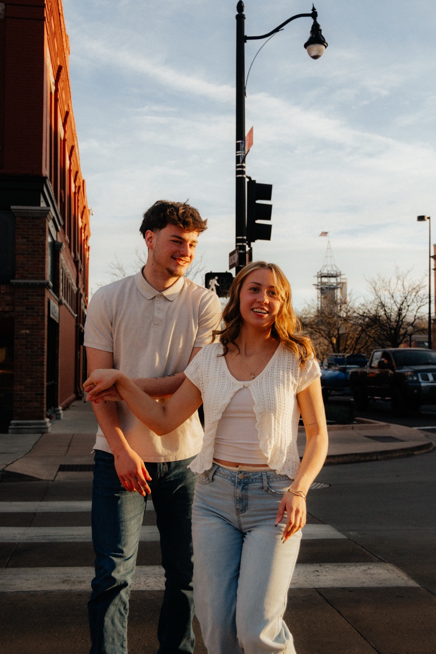 Ellie and Lane walking hand in hand across a sunlit downtown crosswalk