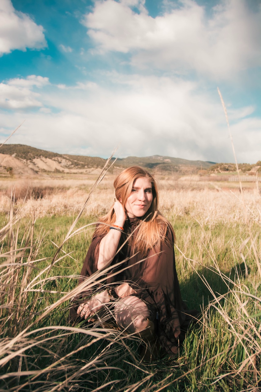 Woman in a brown chiffon dress sitting in tall grass, hand to her hair, smiling softly toward the camera