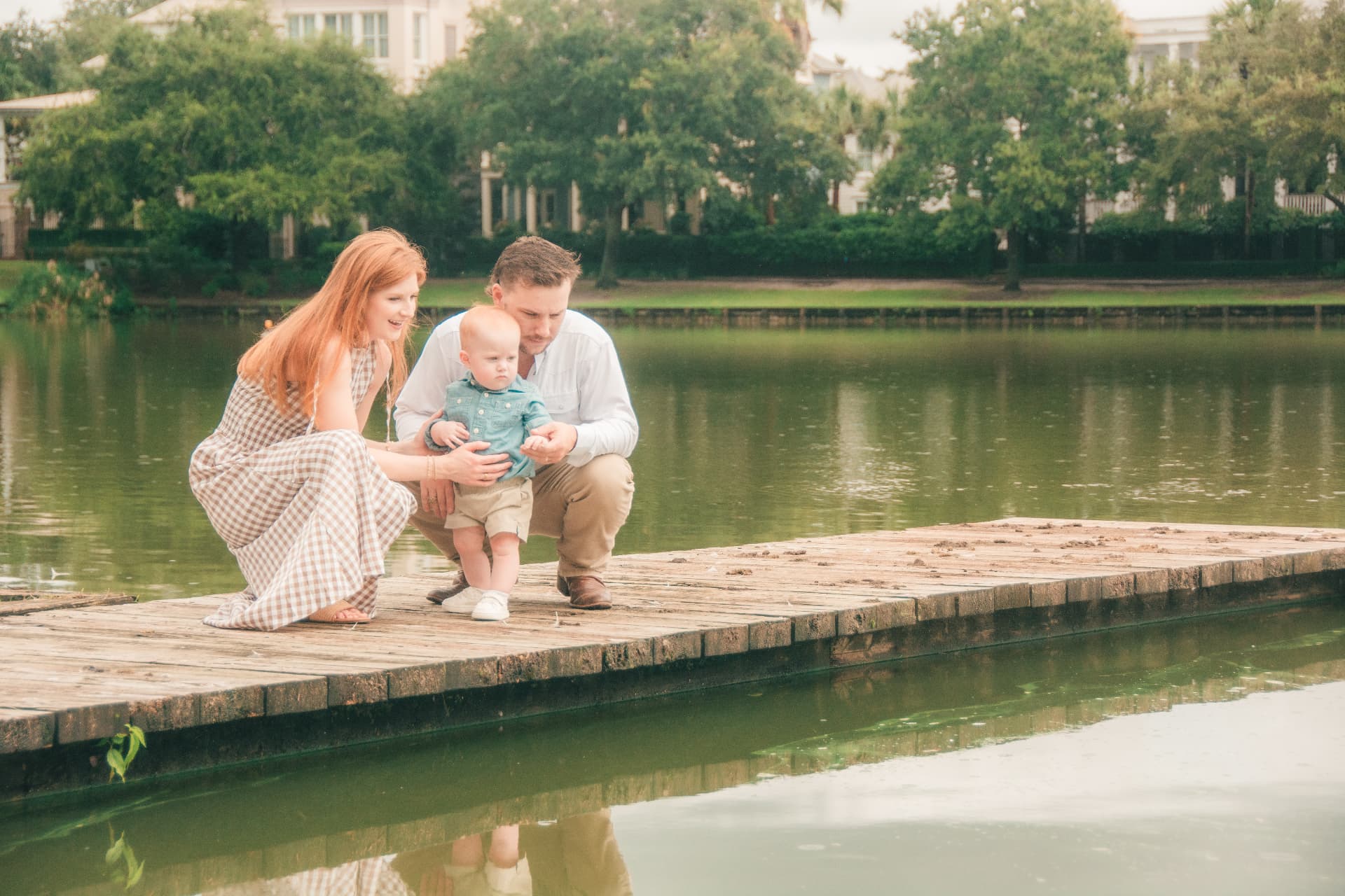 Parents crouching on a weathered wooden dock by the water, holding their baby boy steady as he finds his footing