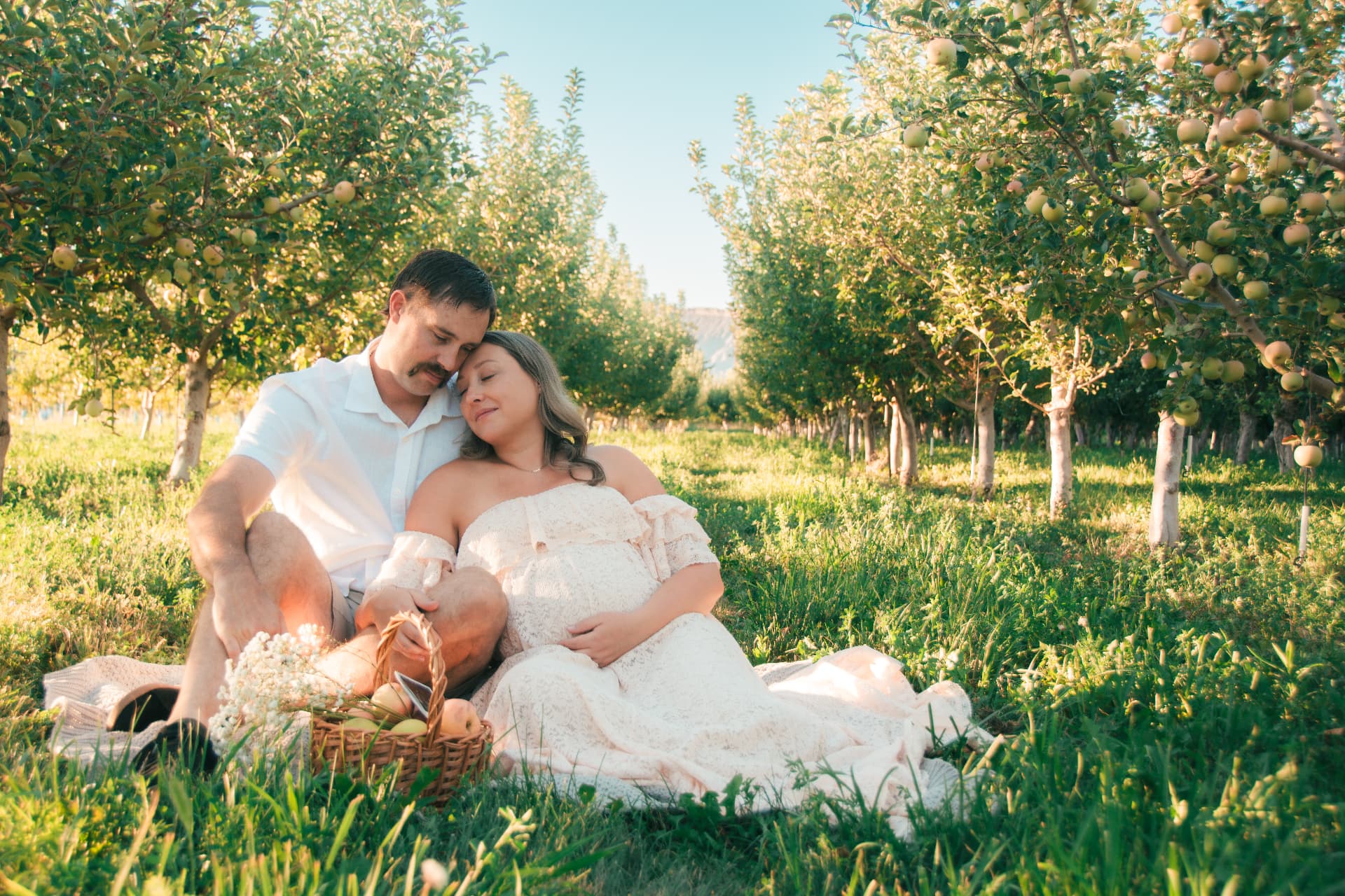 Wide view of the couple sitting on a picnic blanket in the orchard with the mesa behind them