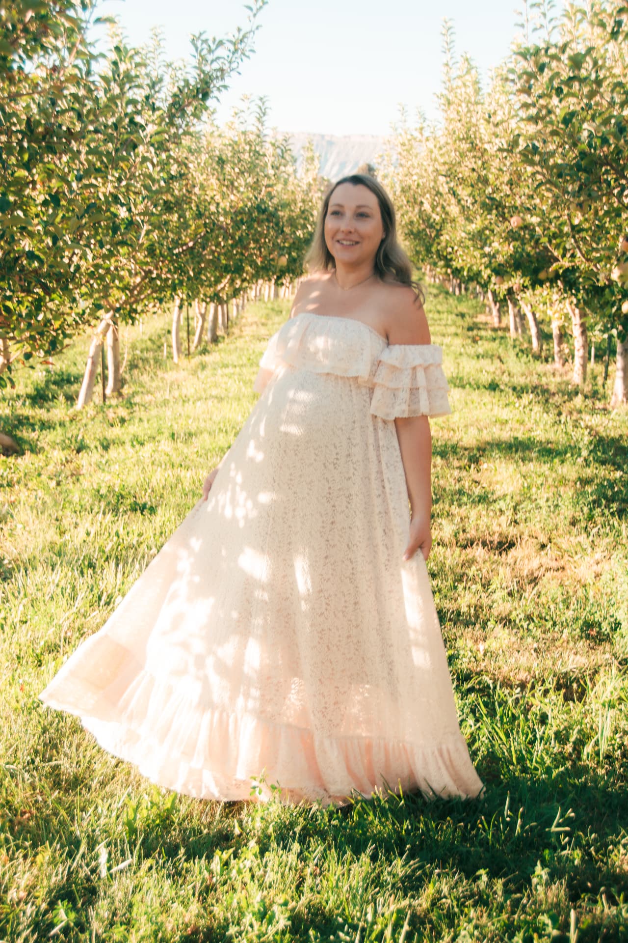 Expecting mother in a peach lace gown standing between rows of apple trees