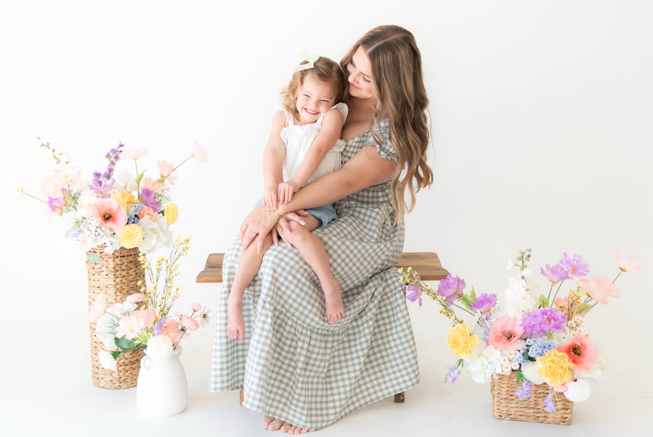 Mom in a sage gingham dress sitting on a wooden bench cradling her toddler daughter, surrounded by spring flowers