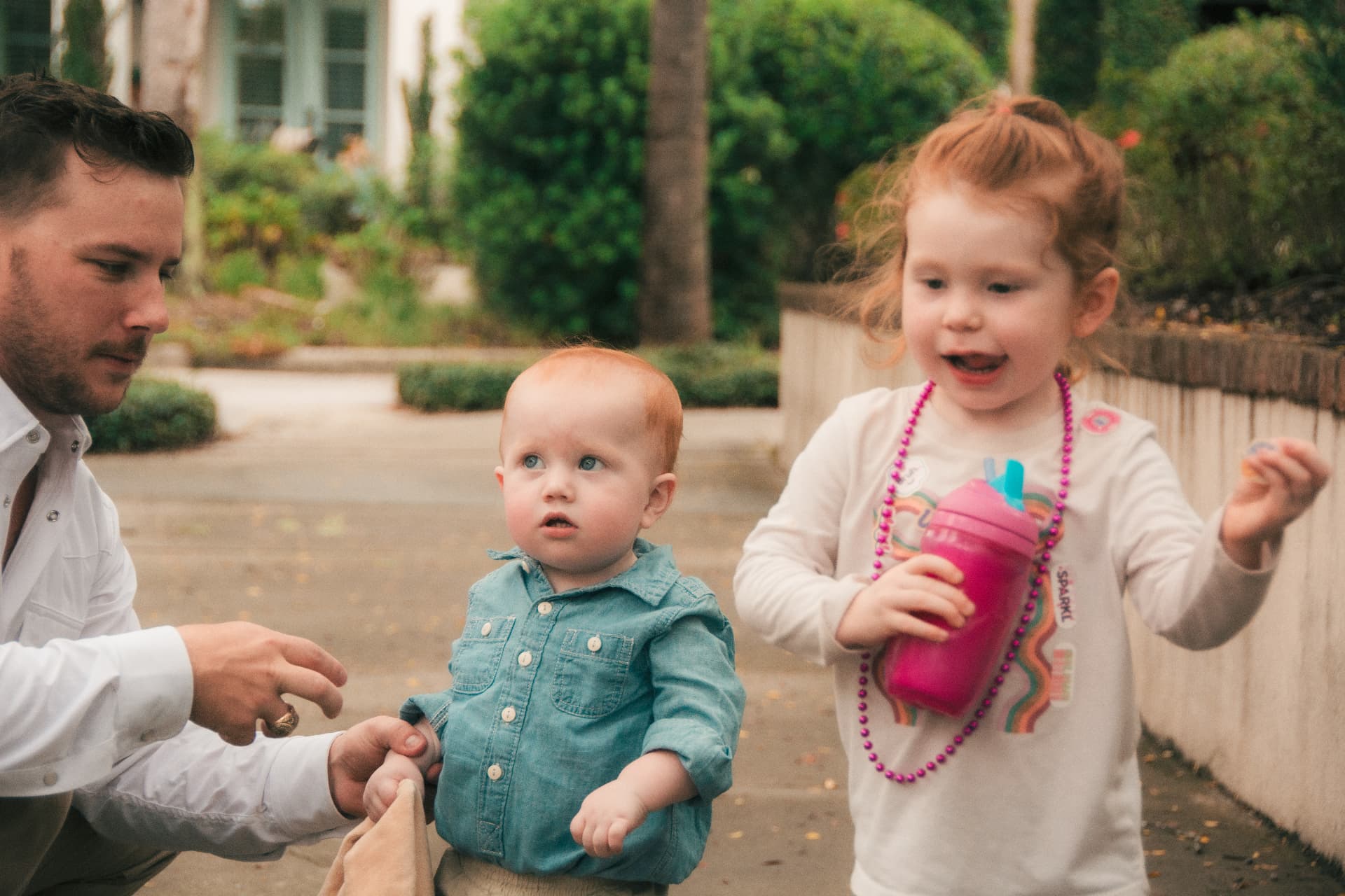 Dad kneeling with the baby while big sister clutches her pink cup beside them