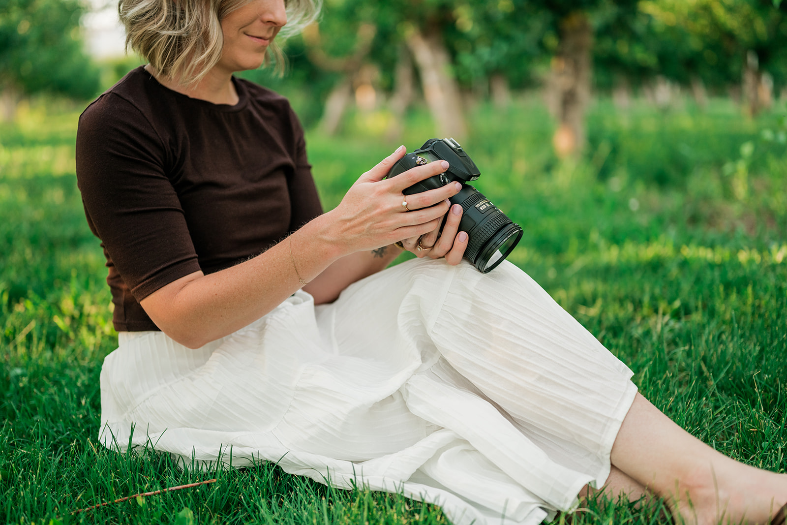 Hannah Hargrave sitting in an orchard holding her camera, wearing a brown top and soft white skirt
