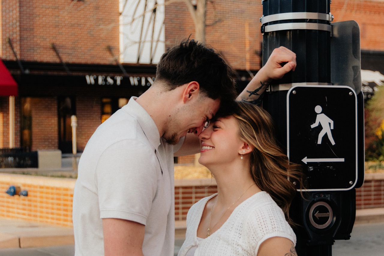 Ellie and Lane laughing forehead-to-forehead beside a pedestrian signal downtown