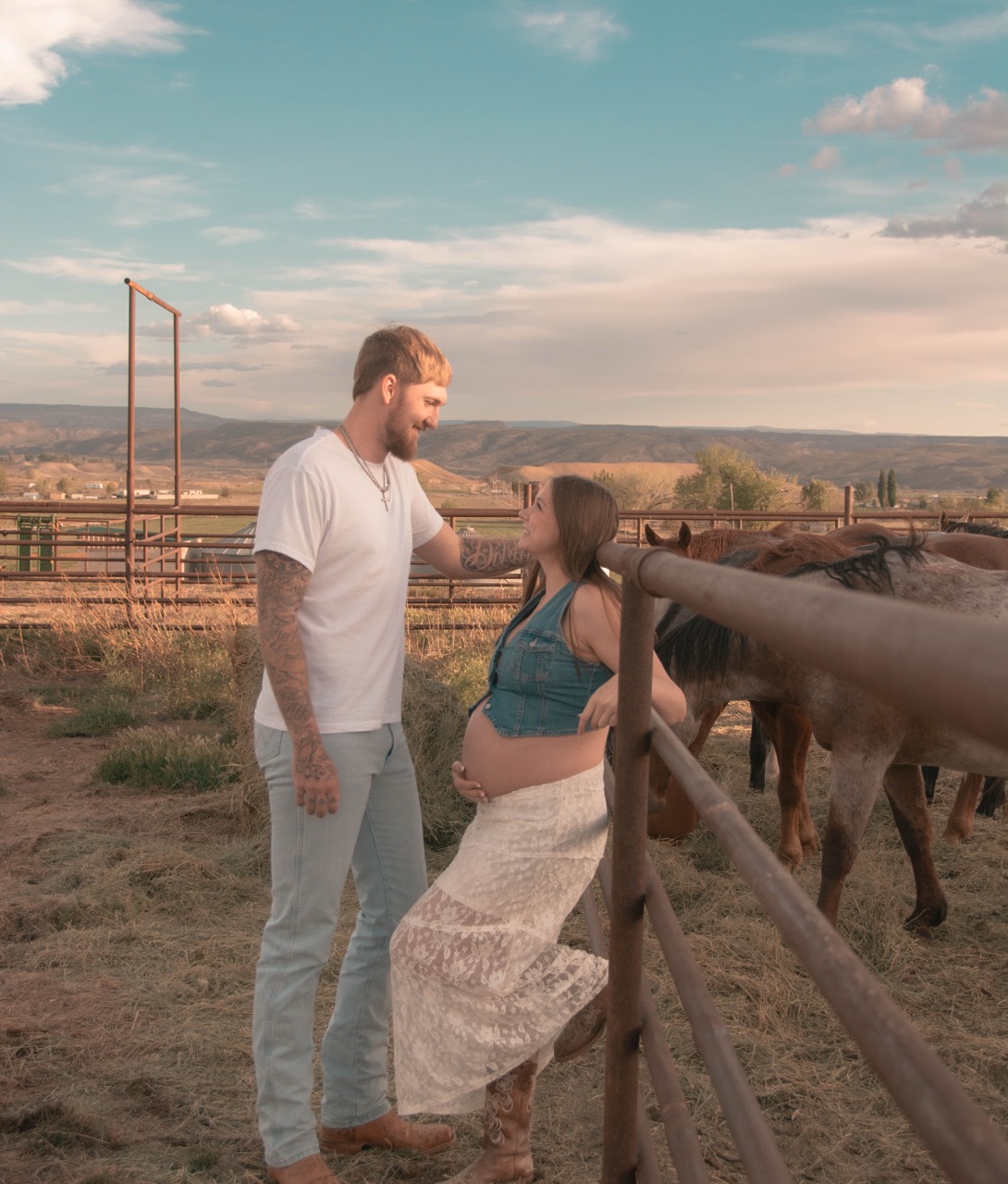 Ashley and Kevin smiling at each other beside the corral fence at golden hour, her hand resting on her pregnant belly