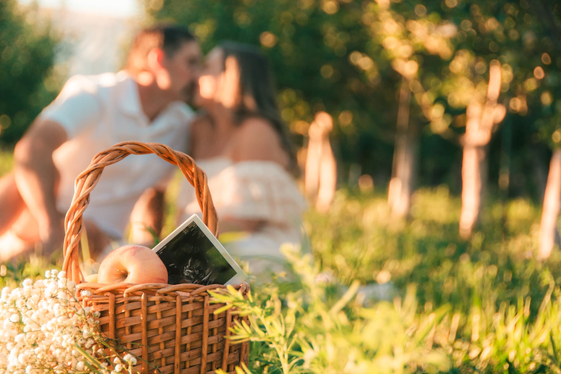 Picnic basket with apples and an ultrasound photo in the foreground as the couple kisses behind it