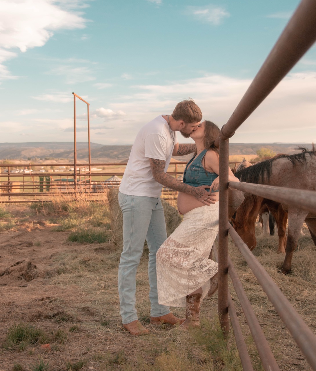 Kevin leaning in to kiss Ashley beside the corral fence as the horses graze behind them