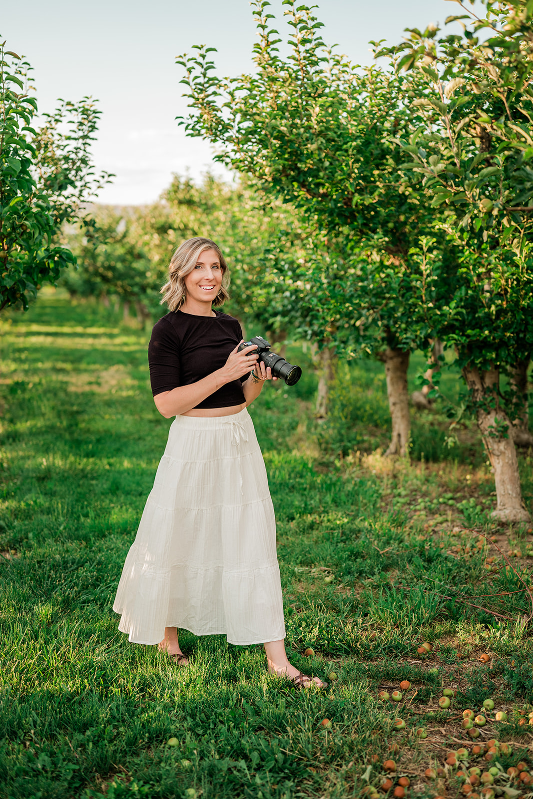 Hannah Hargrave holding a camera in an orchard, wearing a black top and white tiered skirt