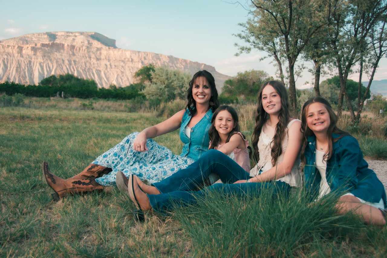 Mother and three daughters sitting in tall grass with cowboy boots and the mesa behind
